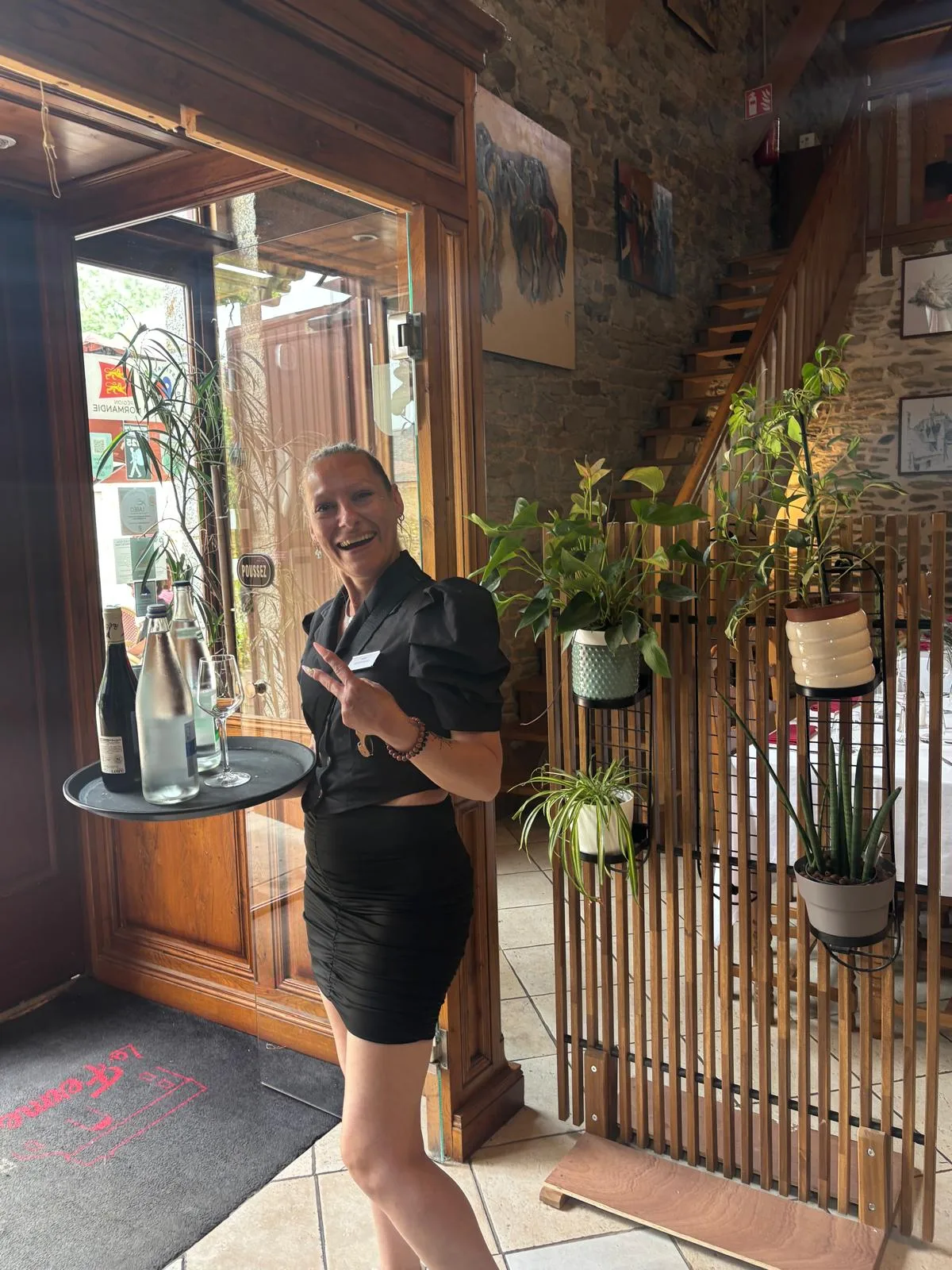 A smiling member of the front-of-house team welcoming guests at the entrance of La Ferme Saint-Michel, beside a tray of wine and bottles